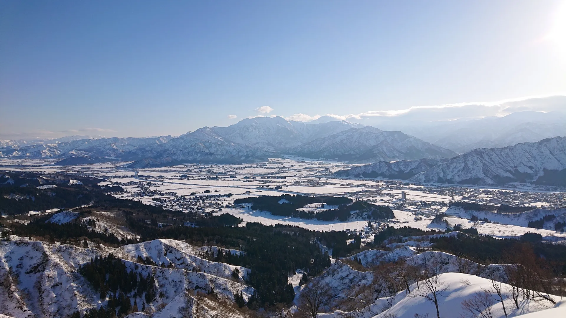 高台から望む冬の魚沼平野。雪におおわれた平野に集落と田畑が広がり、遠景に雪をかぶった山並みが連なる
